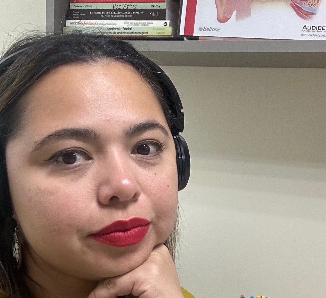 Fga. Mayra Pires com fone de audiometria, diante da estante técnica e do pôster anatômico da orelha